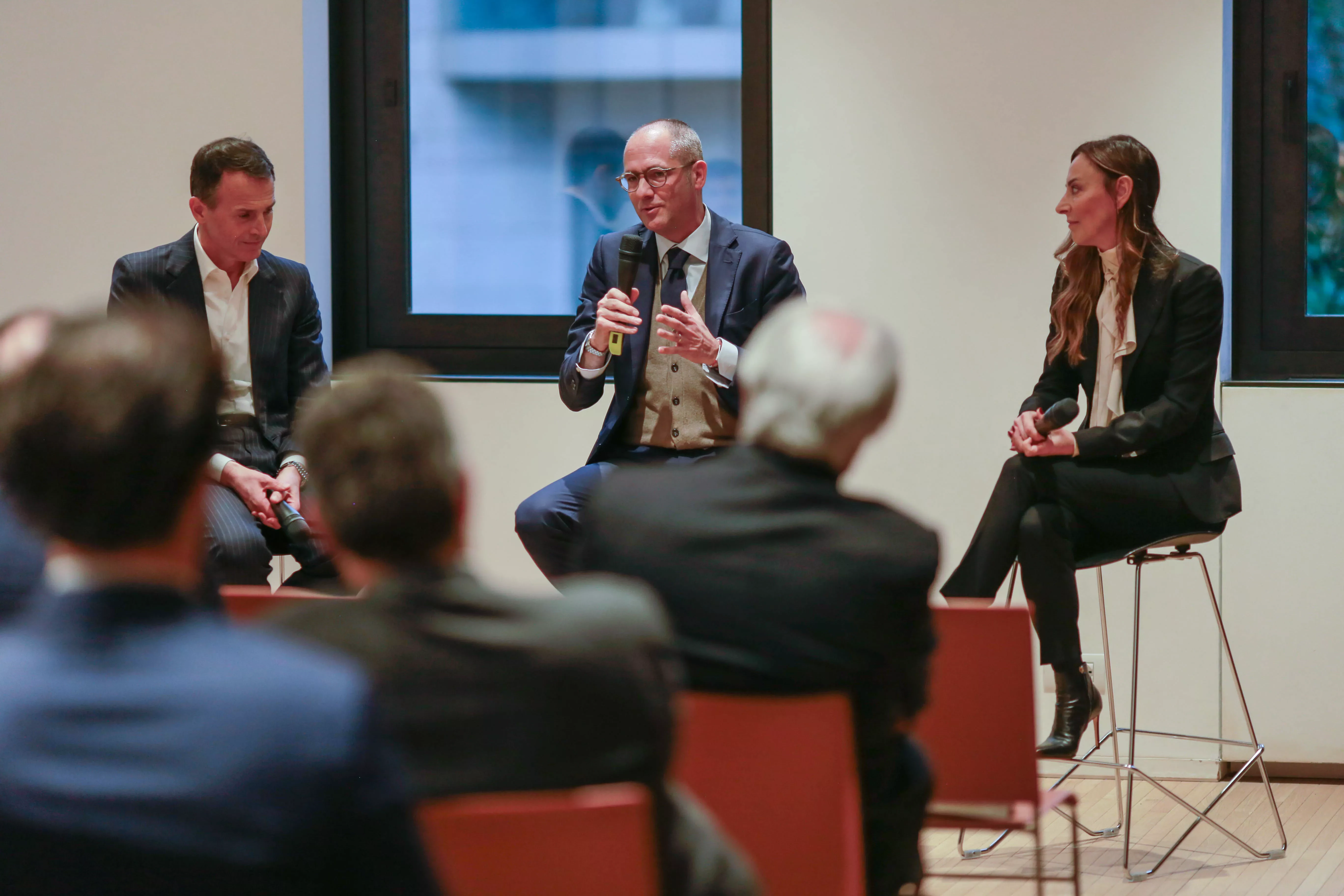 Man in a suit speaks into a microphone while seated on stage with two others during a panel discussion. Audience members are visible in the foreground.