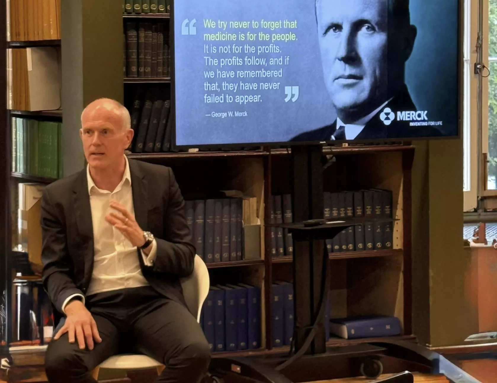 Bald man in a suit speaking while seated in front of bookshelves. A screen beside him shows George W. Merck's portrait and quote about medicine being for the people, not profits.