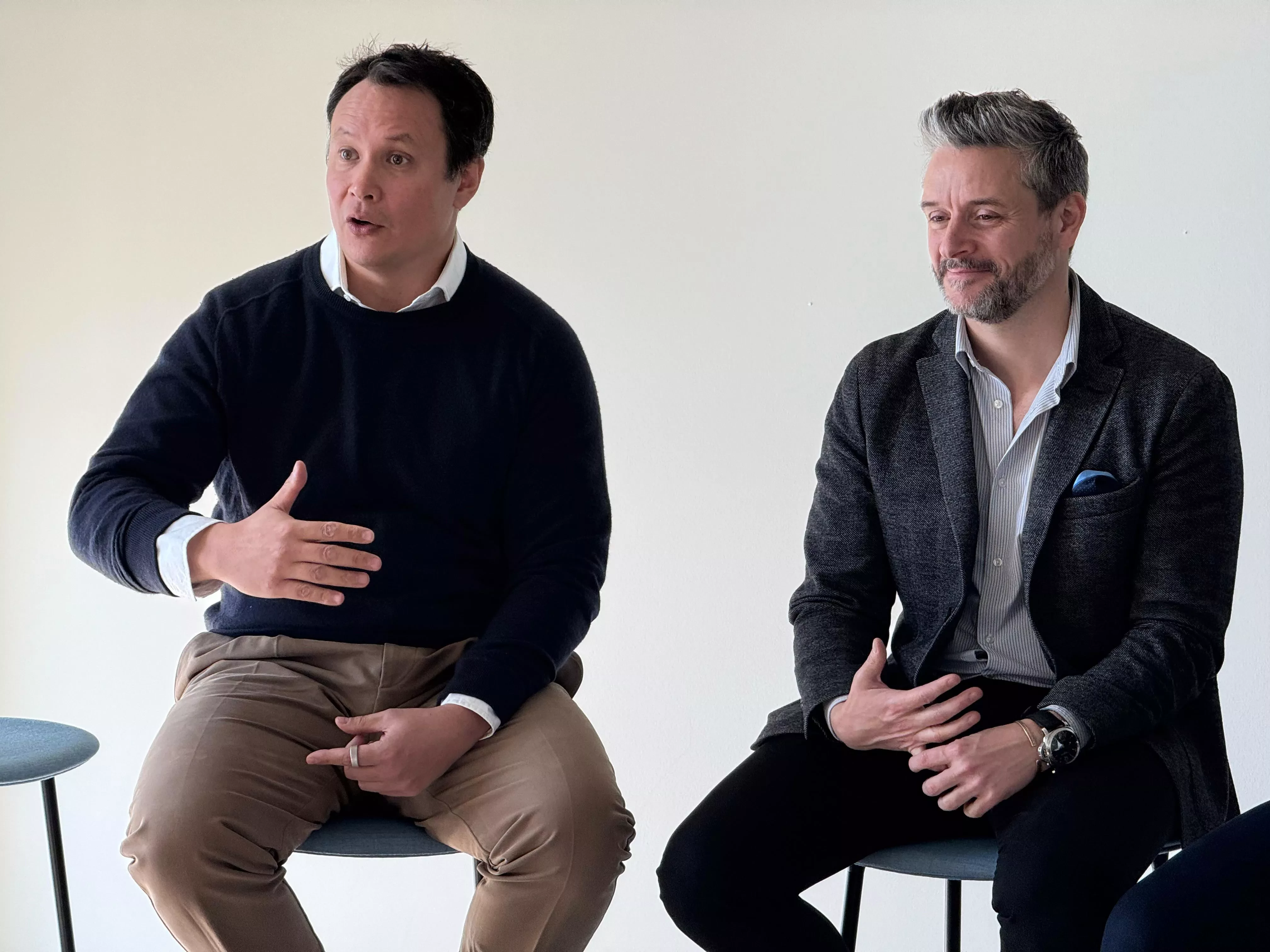 Two men sit side by side during a business talk. The man on the left gestures with his hand while speaking, while the man on the right listens attentively with a slight smile.