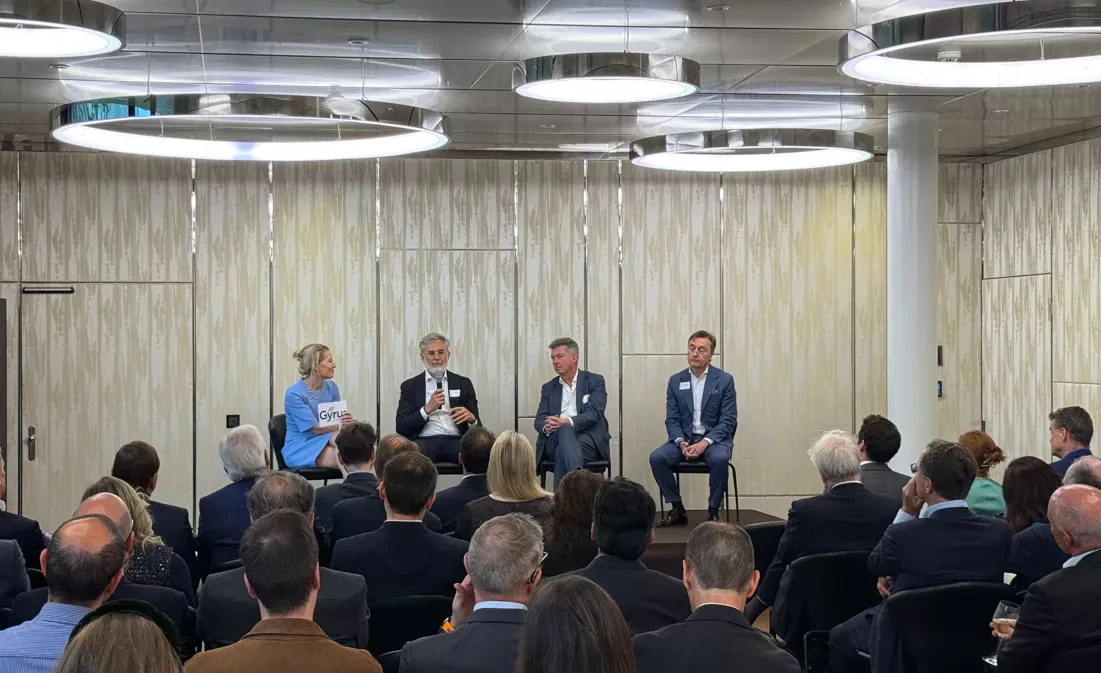 Four speakers sit on stage during a panel discussion in front of an audience. One man speaks while holding a microphone, as the others listen attentively under modern ceiling lights.