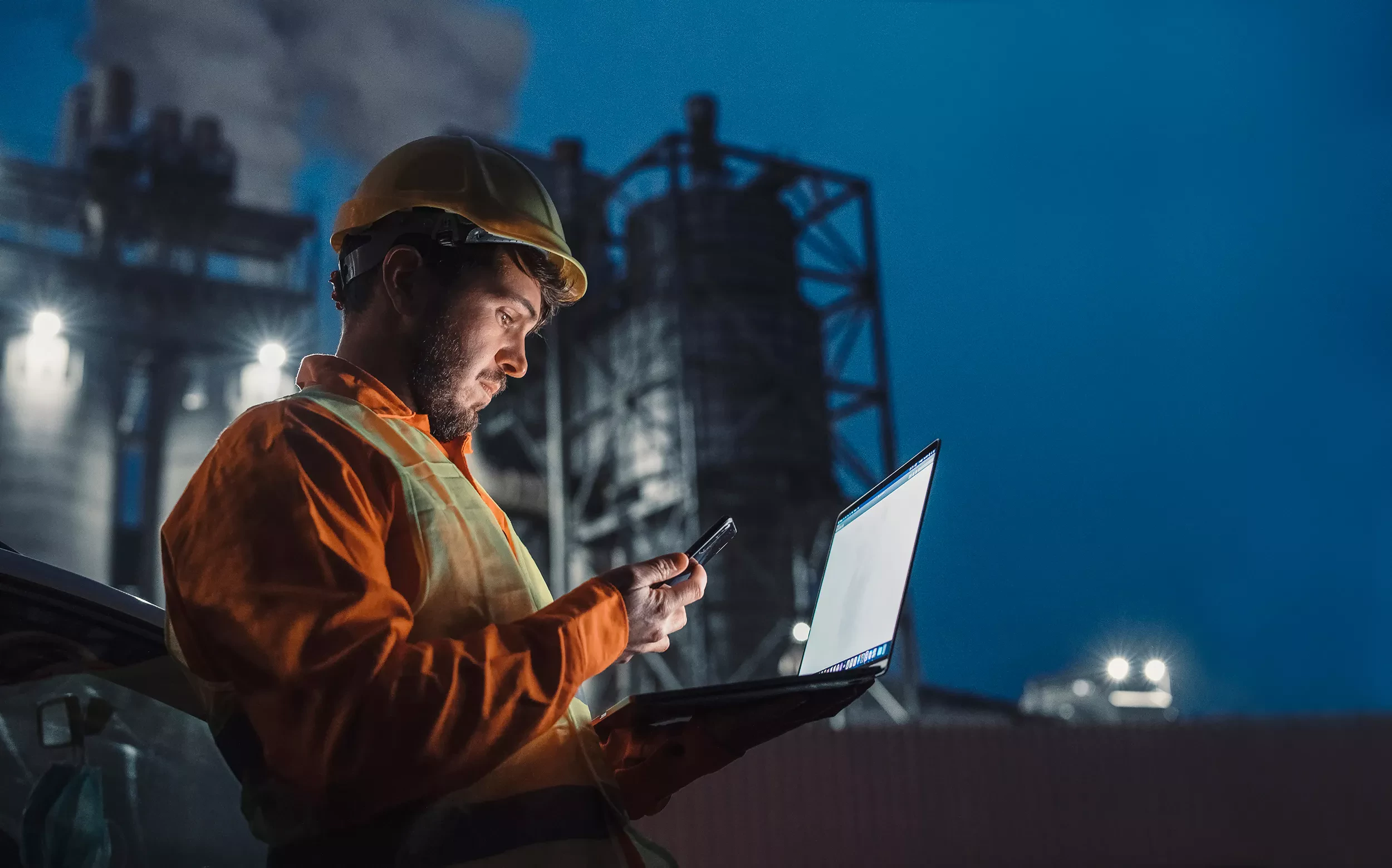 Industrial worker wearing safety gear using a laptop and smartphone at a plant site at night.