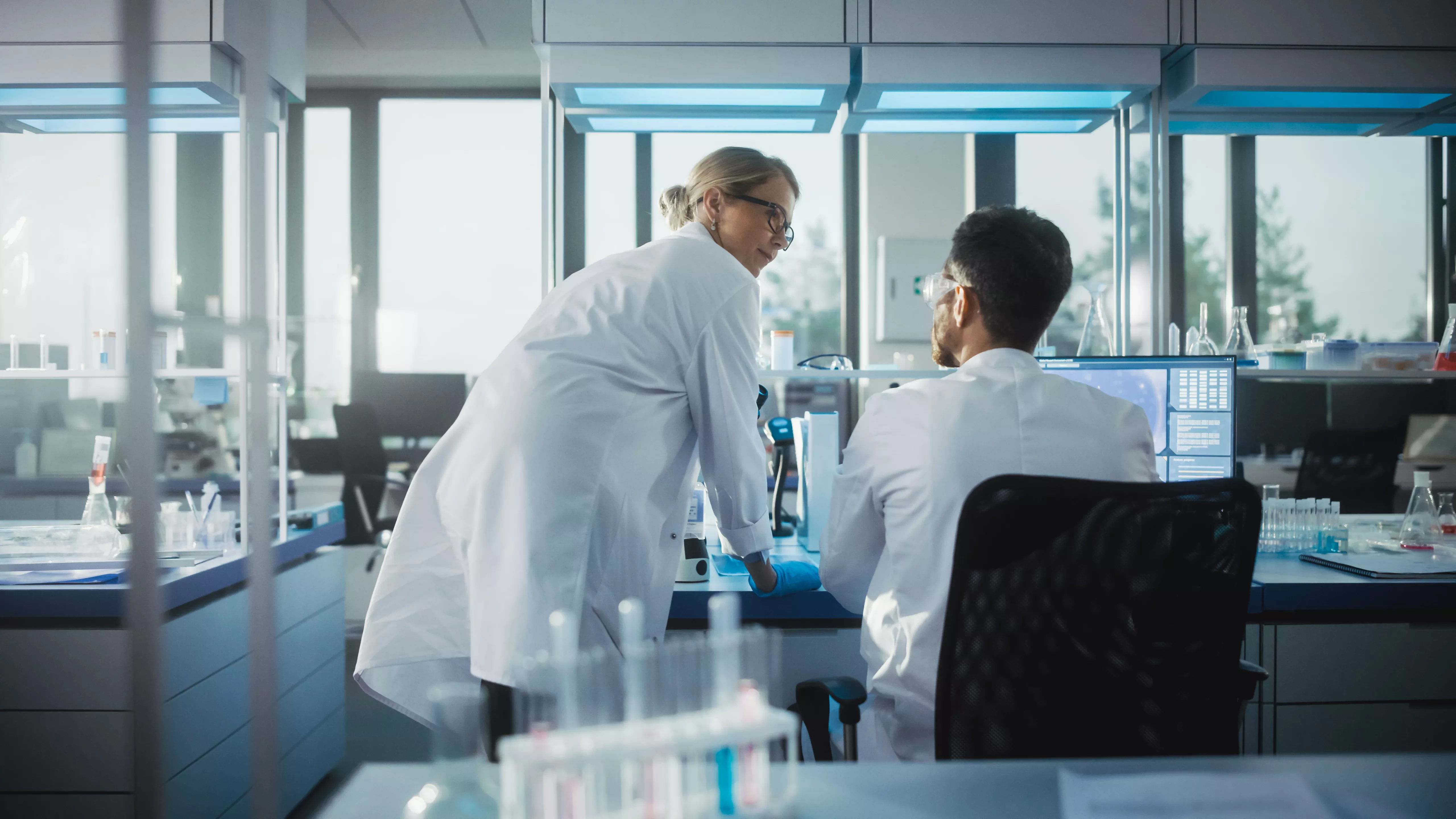 wo scientists in white lab coats discuss work in a modern laboratory, with test tubes and microscopes visible on the bench.