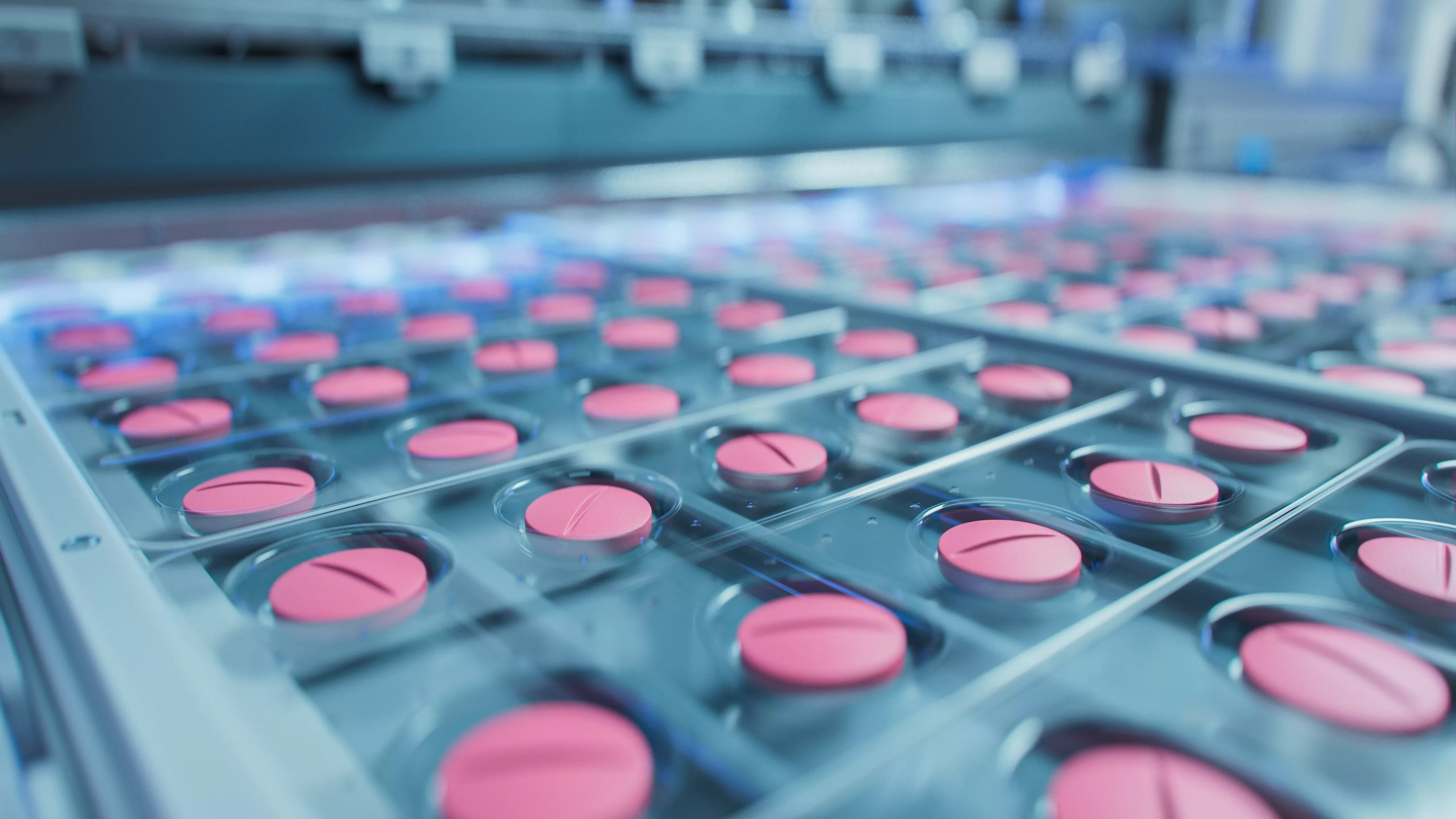 A close-up, high-tech image showing a blister pack of pink, circular pills moving along a pharmaceutical manufacturing or packaging conveyor belt. The scene has a blue, sanitized, industrial tone.