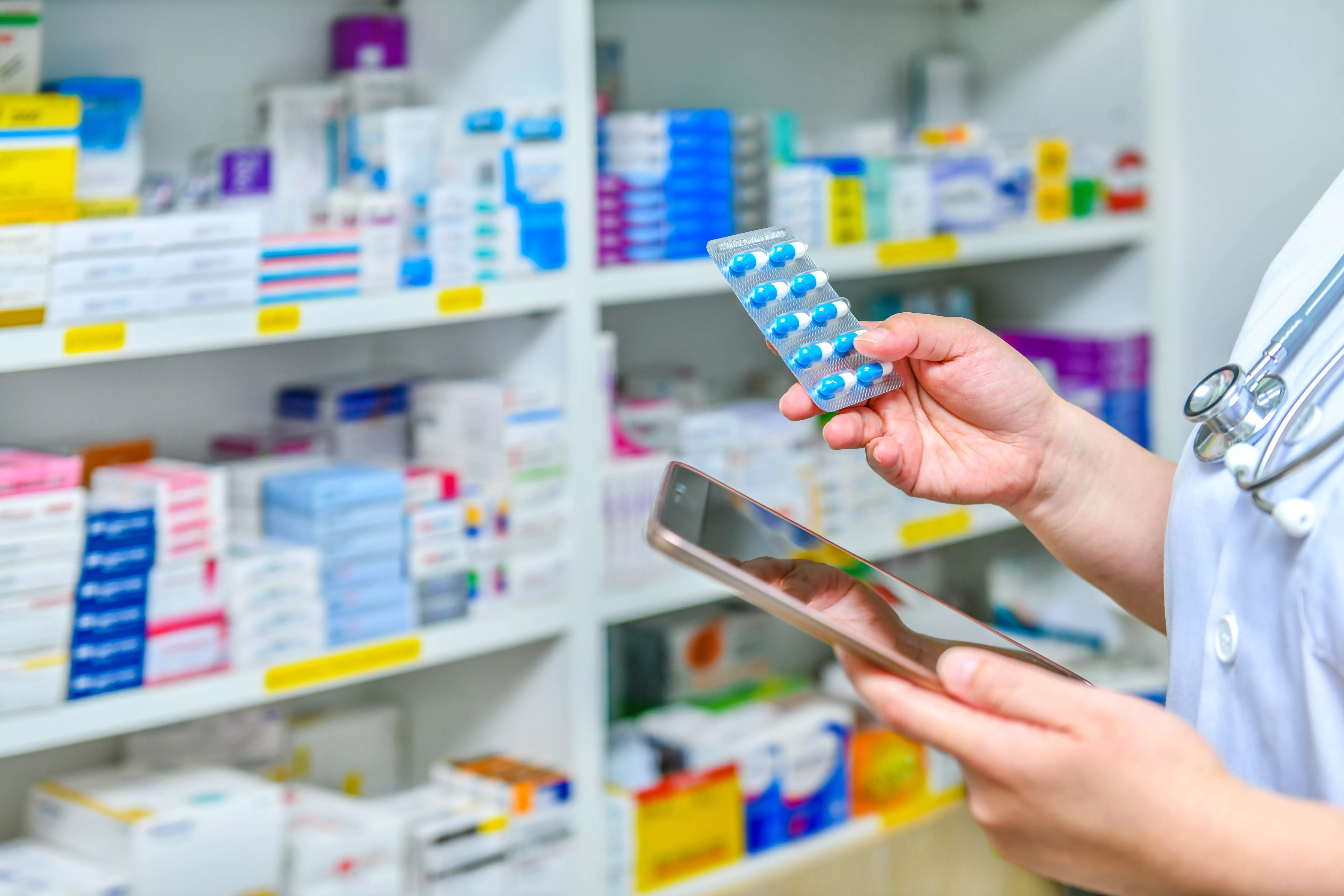 A person in a white coat (likely a pharmacist or doctor) holds a blister pack of blue and white capsules in one hand and a tablet computer/smartphone in the other, standing in front of shelves stocked with various colorful medicine boxes.