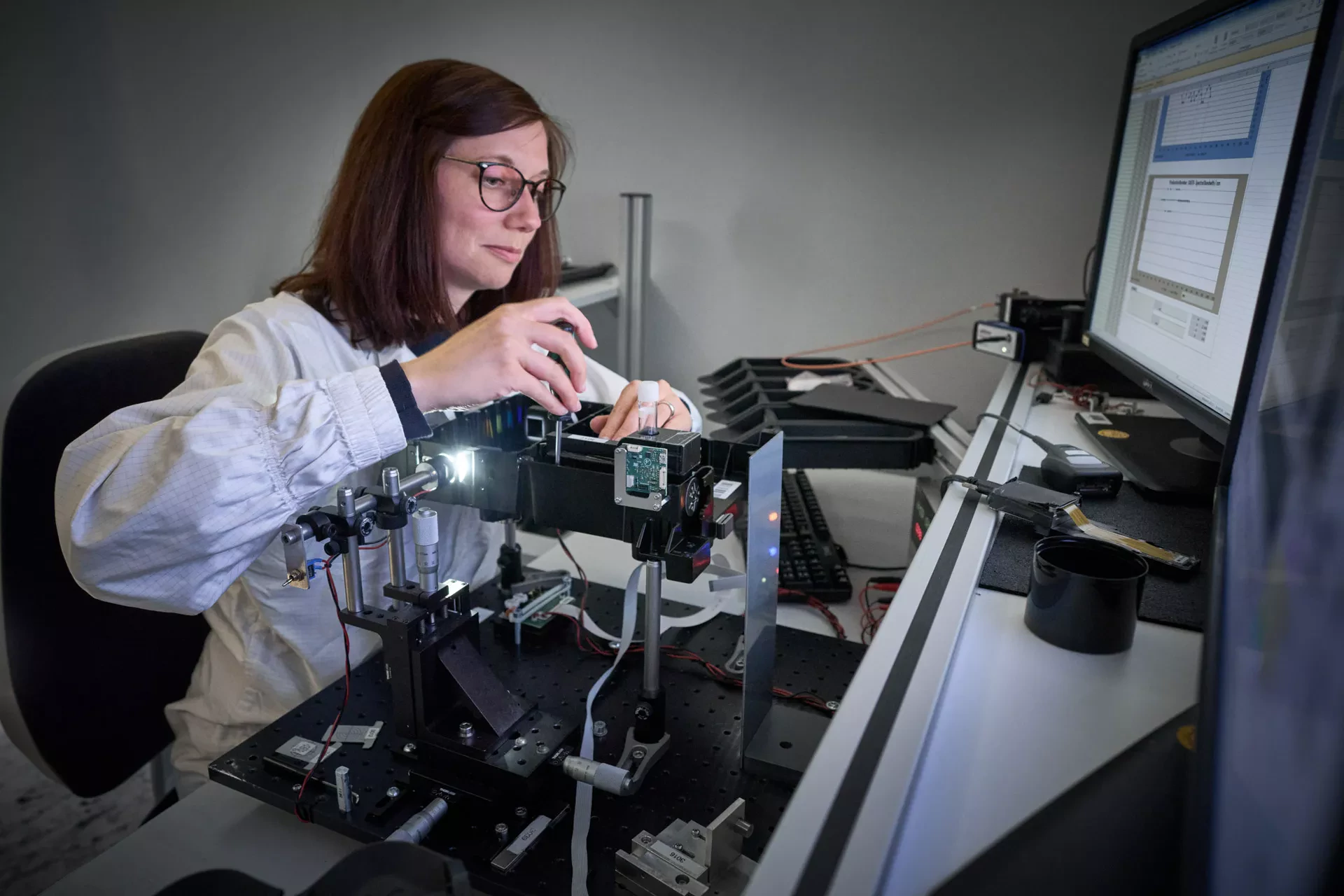 A scientist in a lab coat and glasses precisely adjusts a complex optical system with lasers and sensors on an optical breadboard. Data tables are visible on the adjacent computer screen, indicating ongoing research or calibration.