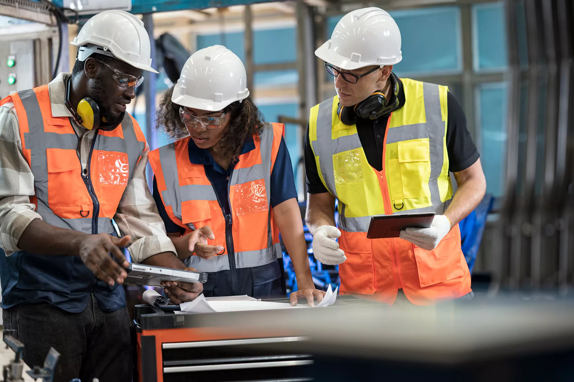  Three workers wearing white hard hats, safety glasses, and high-visibility vests (orange and yellow) are gathered around a table in an industrial setting, reviewing blueprints or documents and discussing data on a digital tablet.