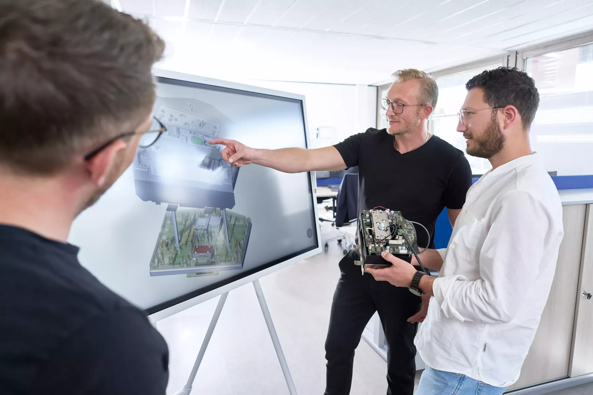  Three colleagues in an office discuss a technical design displayed on a large screen. The man in the center points to the 3D model while holding a complex electronic circuit board prototype.