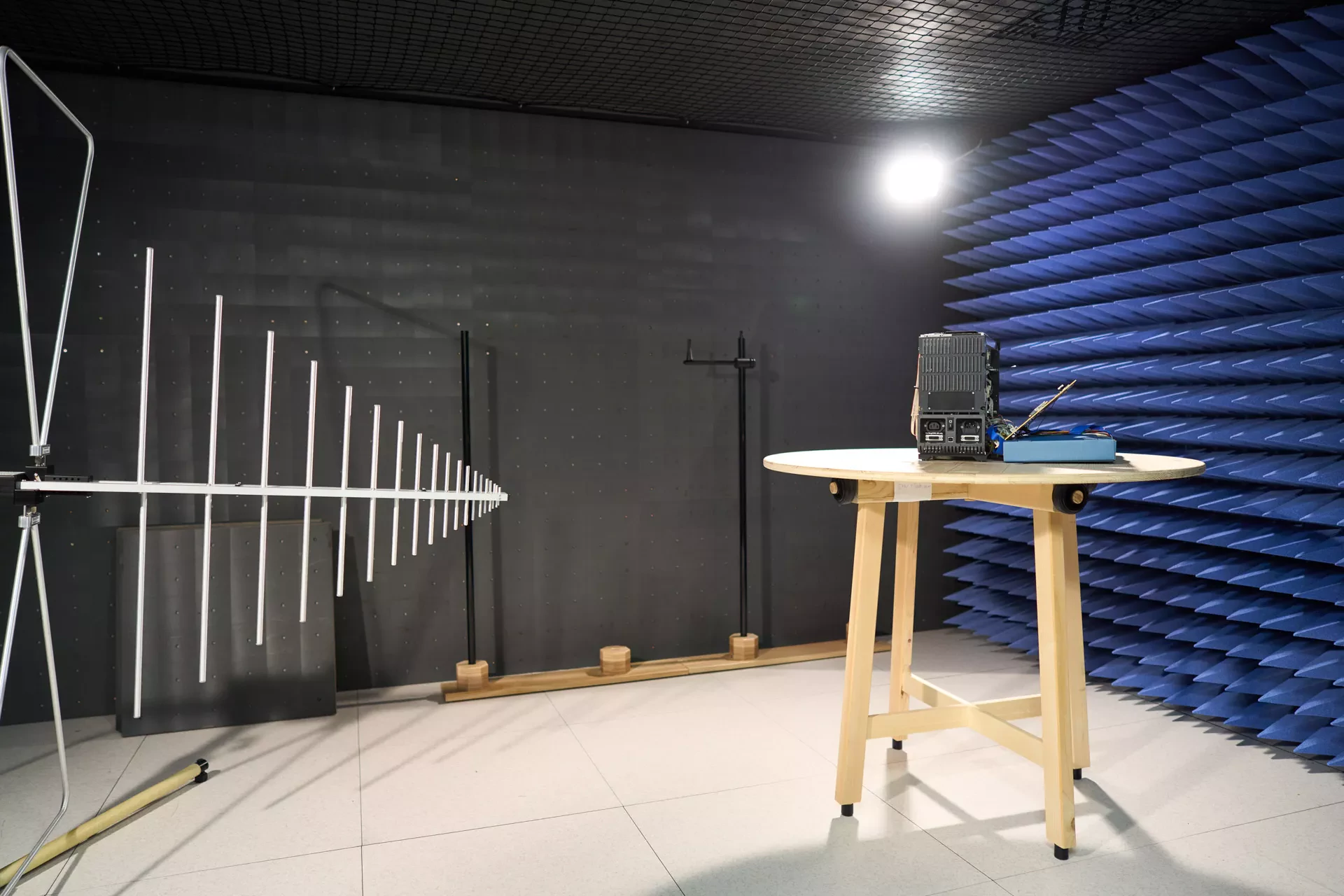 The interior of an EMC (Electromagnetic Compatibility) testing chamber. The room features dark gray walls, blue anechoic foam pyramids on the right, and a large log-periodic antenna on the left. A wooden table holds the electronic device being tested