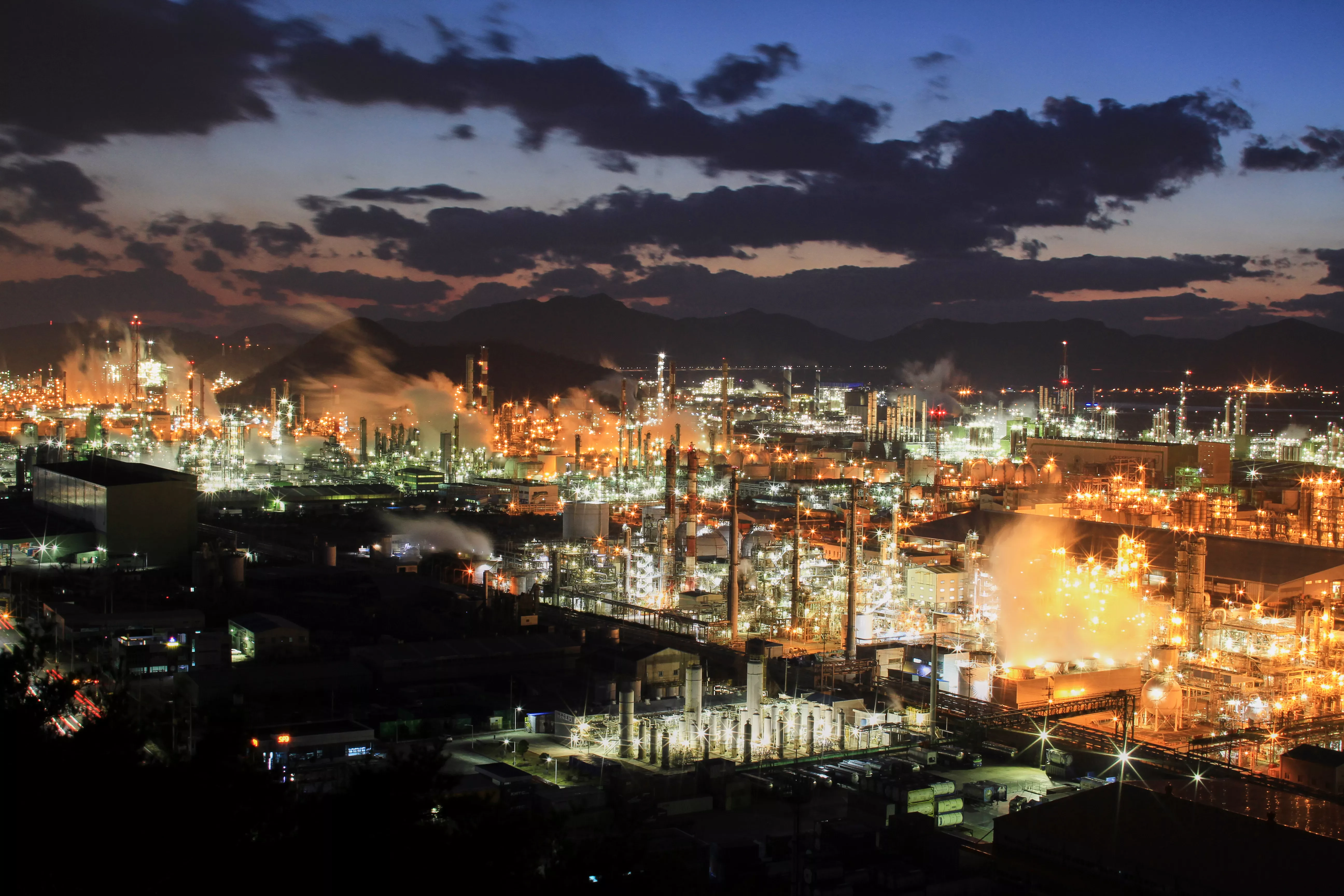 Industrial cityscape at night with brightly lit factories and smokestacks emitting steam, symbolizing sustainability challenges addressed by DuPont Sustainable Solutions’ acquisition of Sofies Group.