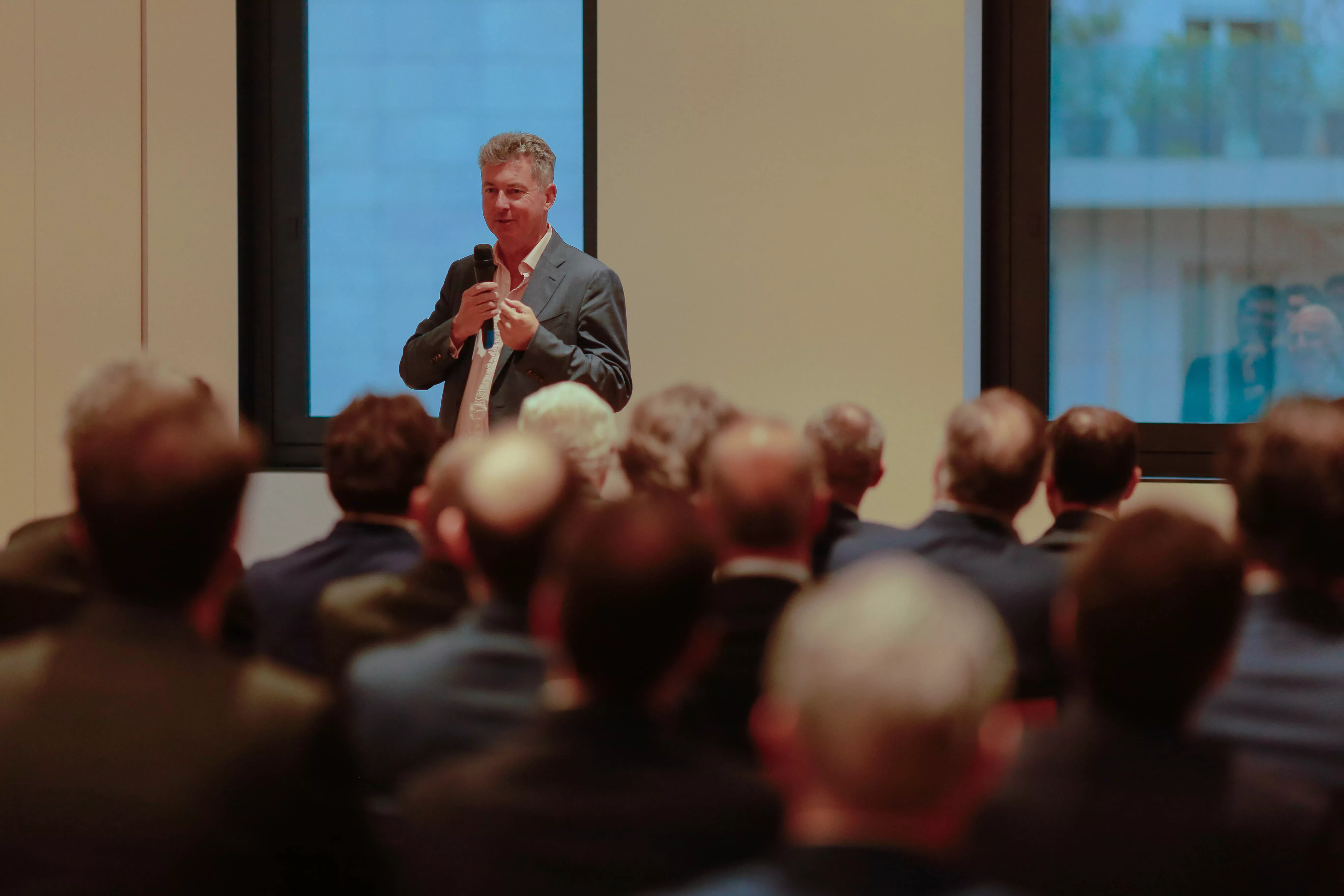 Man in a suit speaks into a microphone during a Gyrus Capital event, addressing an audience seated in a conference room.