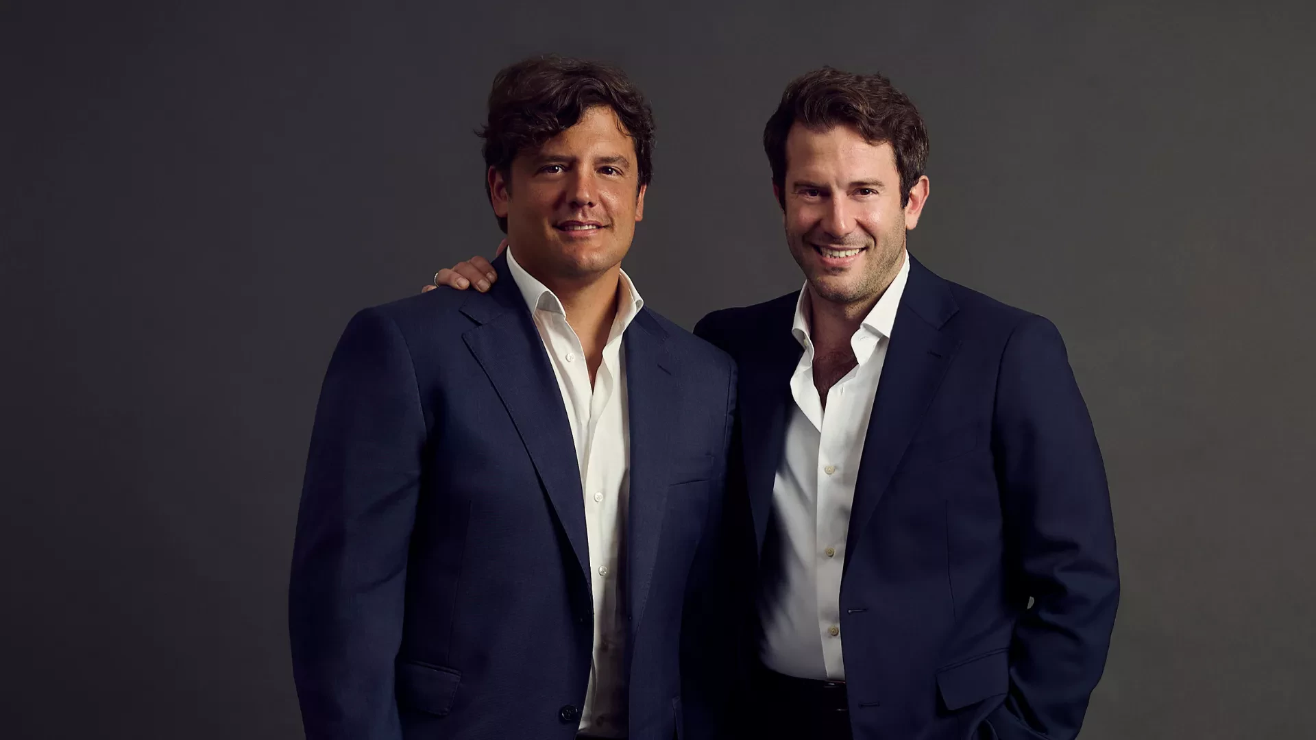 A portrait photo of two men in dark blue suits and white shirts, smiling and standing shoulder-to-shoulder against a solid dark gray background. The man on the left has his right arm resting on the shoulder of the man on the right.