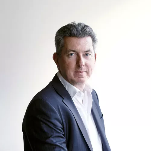 Portrait of a middle-aged man with short gray hair, wearing a dark suit and white shirt, standing against a plain light background. He looks directly at the camera with a composed and confident expression.