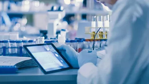 Scientist in lab coat and gloves working with a tablet next to test tubes, symbolizing Essential Pharma’s focus on niche medicines and innovation in healthcare research.