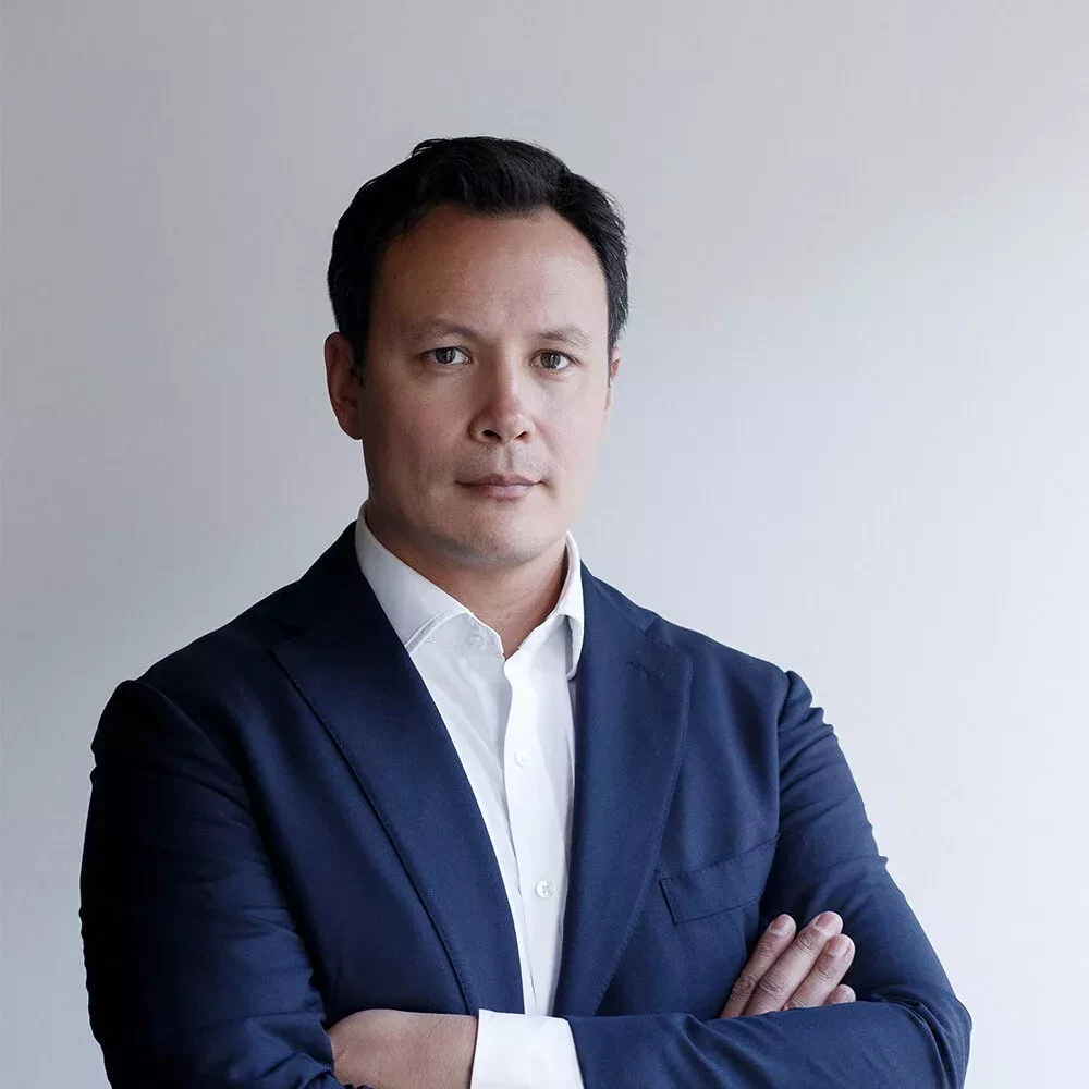 Portrait of a man with short dark hair, wearing a dark blue suit and a white shirt, standing with arms crossed. He looks directly at the camera with a serious and composed expression, set against a plain light background