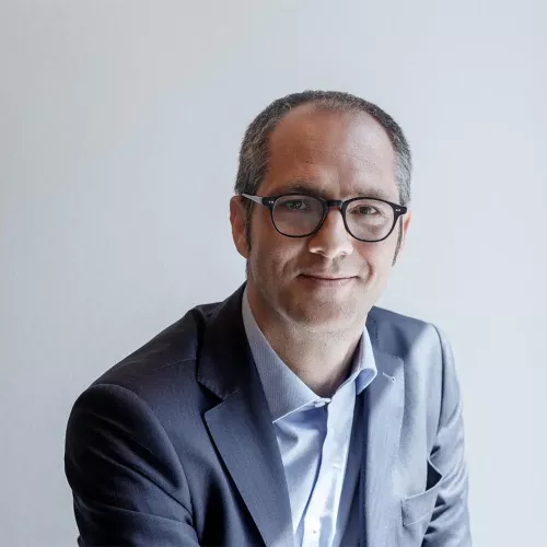 Portrait of a man with short hair and glasses, wearing a dark suit and light blue shirt. He is slightly smiling and looking directly at the camera, set against a plain light background.