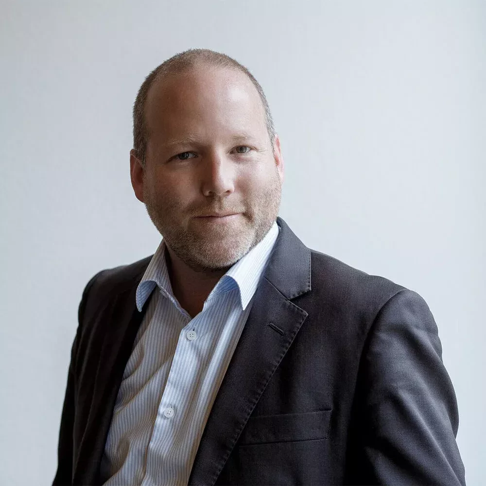 Portrait of a man with short light hair and a beard, wearing a dark suit jacket over a light blue striped shirt. He has a slight smile and is looking confidently at the camera, set against a plain light background.