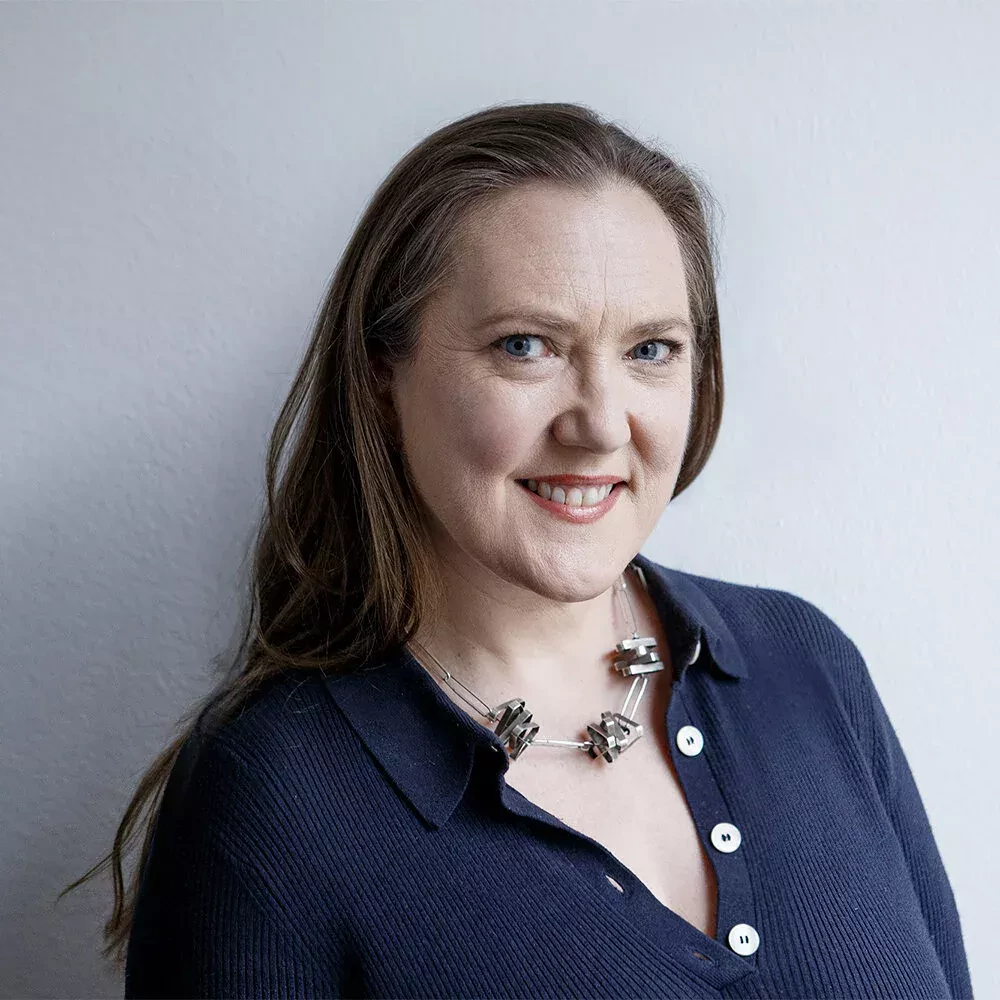 Portrait of a woman with long straight brown hair, wearing a dark blue buttoned shirt and a chunky silver necklace. She is smiling warmly and looking at the camera, set against a plain light background.