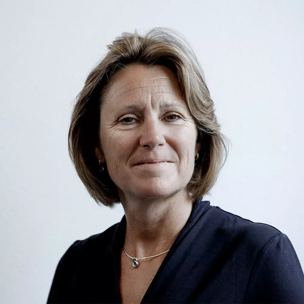 Portrait of a woman with short light brown hair, wearing a dark blouse and a delicate necklace with a small pendant. She has a calm, confident expression and is photographed against a plain light background.