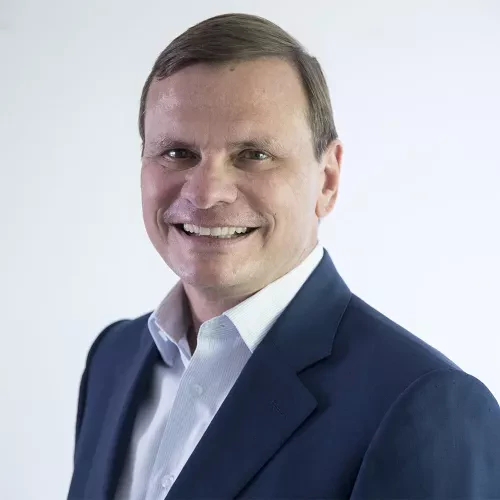 Portrait of a man with short light brown hair, wearing a white shirt and a dark blue blazer. He is smiling warmly and facing the camera, with a light plain background.